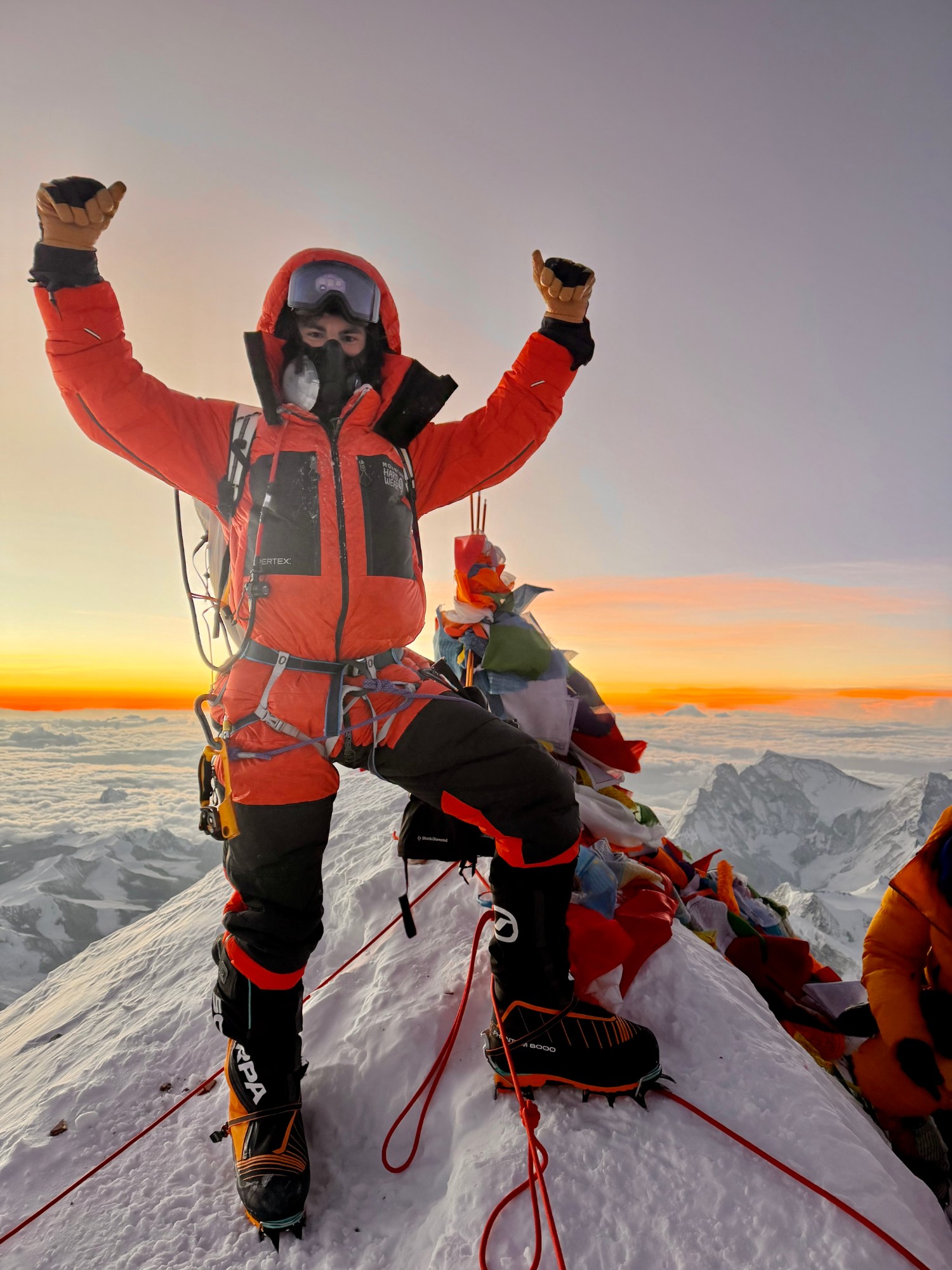 James Jackson on the summit of Everest at sunrise, arms raised beside the prayer flags
