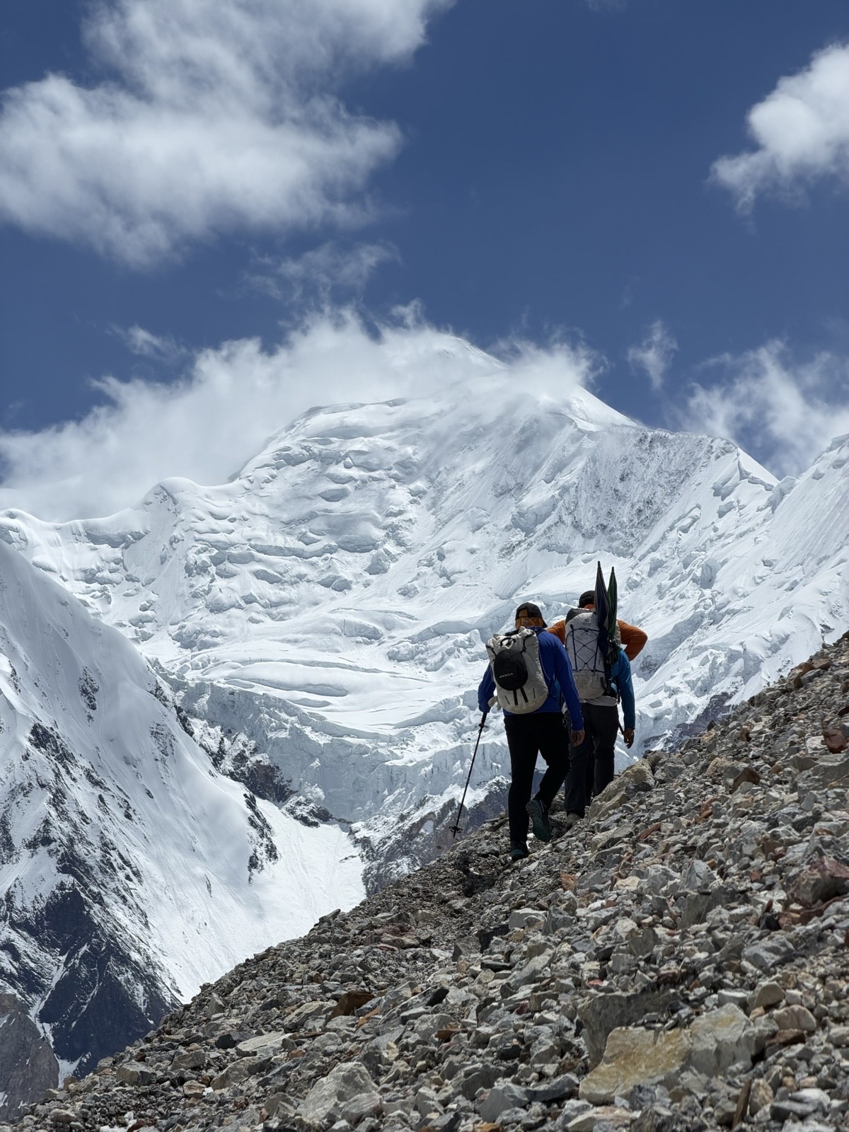 Climbers crossing a rocky slope below the snow covered peaks of the Gasherbrum range